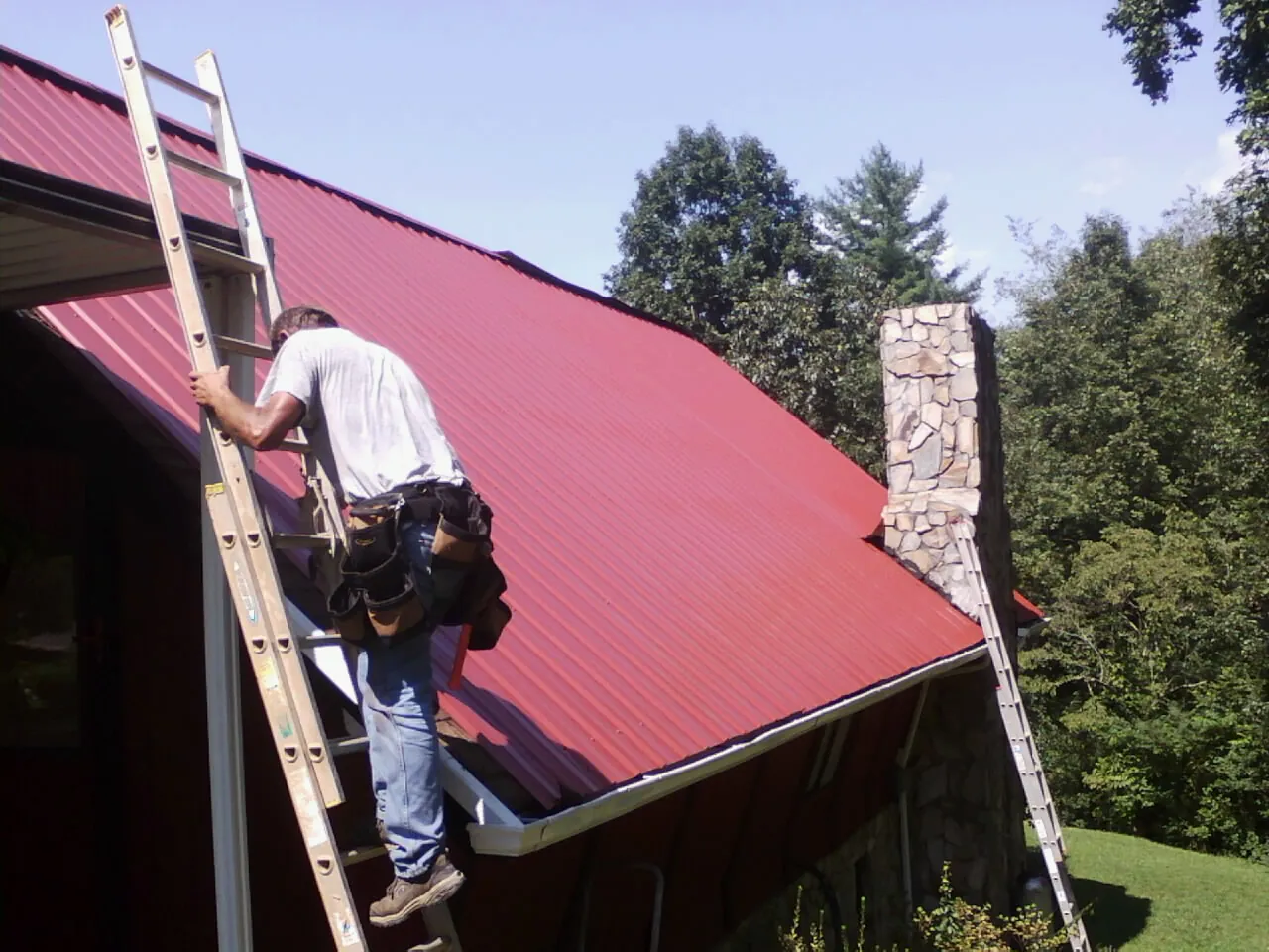 Roofer on a ladder beside a red metal roof and stone chimney — Soffit & Fascia Repair by Roofing Brothers in Summerland